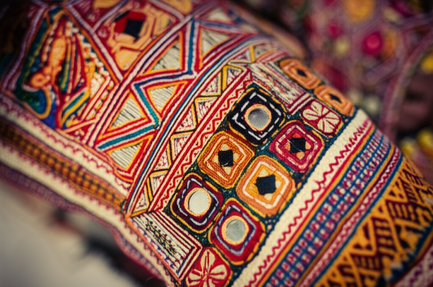 alt_text: Close-up of vibrant, intricately stitched Banjara embroidery. Rich reds, yellows, blues, and greens. Vintage, slightly faded look.